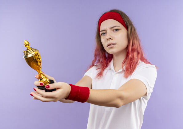 头带戴着头巾 表情严肃地拿着奖杯的年轻运动女郎站在紫色的墙上女人女人人