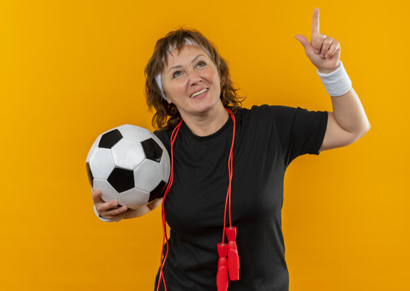 目录中年运动女性 身穿黑色t恤 头带 手持足球 食指朝上 微笑着站在橙色的墙上人姿势球