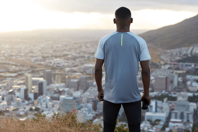 教练黑色运动男的后视图穿着休闲t恤 拿着装有新鲜饮料的瓶子 俯瞰城市建筑 欣赏山景 享受速度和户外锻炼运动概念全景运动员山