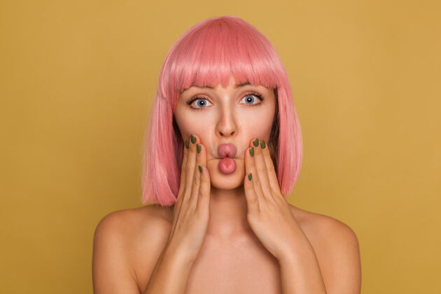 肩膀年轻漂亮的睁着眼睛的女人 粉色的鲍伯发型 双手举在脸颊上 站在芥末墙上做鬼脸姿势爱手