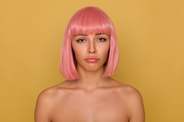 站一头短发的年轻漂亮的粉红色头发的女性 哭着看着镜头 撅着嘴 光着肩膀站在芥末墙上心情肖像年轻