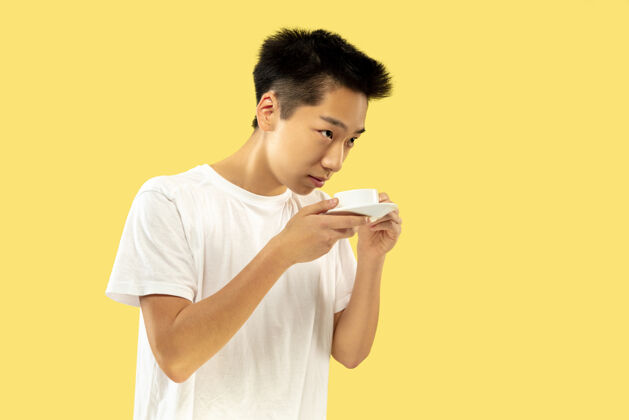 高管黄色摄影棚背景上的韩国年轻人半身像穿白衬衫的男模喝咖啡 感觉快乐人类情感的概念 面部表情正面视图流行色彩男士肖像亚洲人