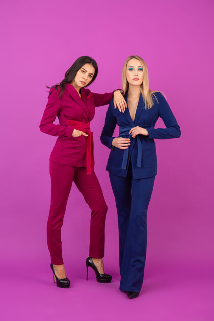 人时尚风格两位微笑迷人的女士在紫罗兰色的墙壁上穿着紫色和蓝色的时尚多彩的晚礼服 春天的时尚潮流夹克时尚优雅