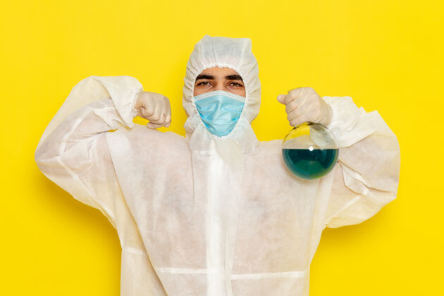保护正面图穿着特殊防护服的男科学工作者拿着黄色表面弯曲的蓝色溶液烧瓶危险面罩服装