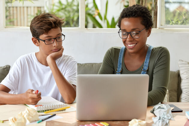 表达团队合作的理念黑人聪明的女老师和她的学生一起看培训视频 集中到笔记本电脑 连接到互联网 坐在桌面上用记事本做记录内容眼镜项目