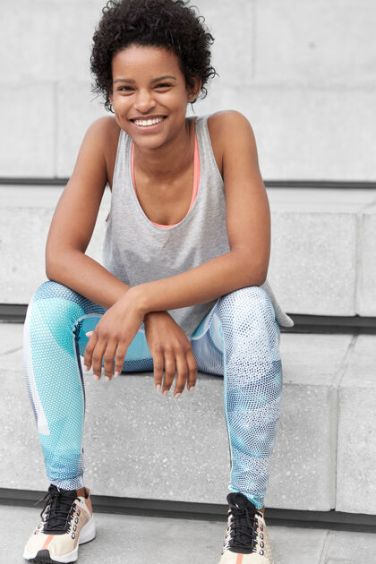 瘦身照片中 自信的黑人女士穿着运动服 表情轻松愉快 笑容灿烂 乐于赢得体育比赛 坐在楼梯旁 准备实现新的目标种族 运动动作上衣背心