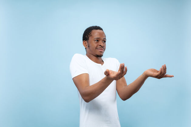 面部在蓝色工作室背景上 年轻的非洲裔美国男模穿着白衬衫的半身特写肖像人类的情感 面部表情 广告概念怀疑 询问 显示不确定性商人就业长相