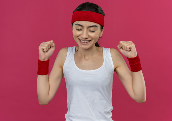 头带身穿运动服 头箍紧握拳头的年轻健身女士站在粉红色的墙上为自己的成功感到高兴和兴奋成功兴奋快乐