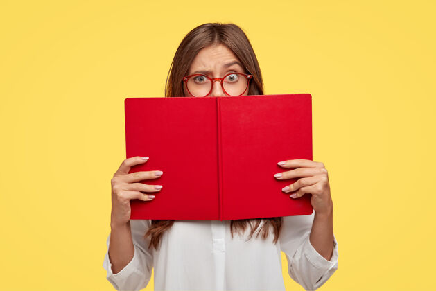 学习迷惑不解的黑发女人扬起眉毛 困惑地看着红色的教科书 穿着白衬衫 靠着黄色的墙壁做模特 学习新的信息 准备家庭作业 读故事站立高加索室内