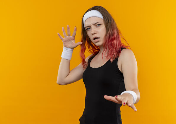人身穿运动服的年轻健身女士伸出手掌 站在橙色的墙上做着一个带着恐惧表情的防御手势年轻人制作手势