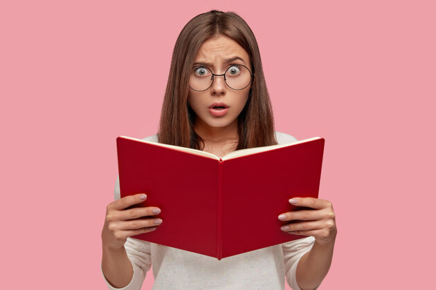 青少年情绪惊讶的欧洲女人拿着课本 有害怕的表情 在通过入学考试前担心 戴着圆眼镜隔着粉红色的墙红皮书的女人女孩大学学校震惊