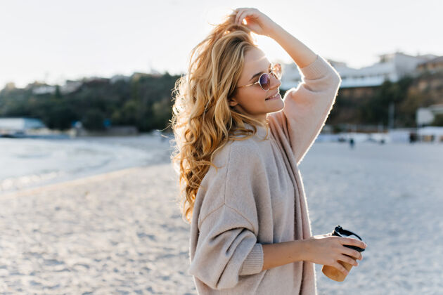 休息穿着米色超大号毛衣 戴着棕色太阳镜的时尚美女blinde带着一杯硬纸板茶走在沙滩上乐趣自然茶