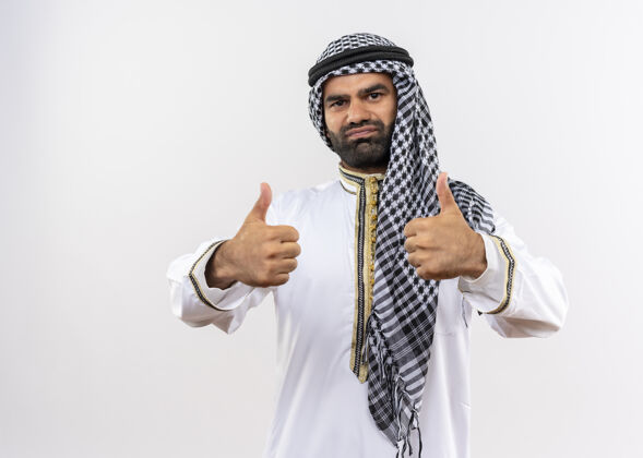 站身着传统服装的阿拉伯男子微笑着竖起大拇指站在白墙上拇指穿阿拉伯语