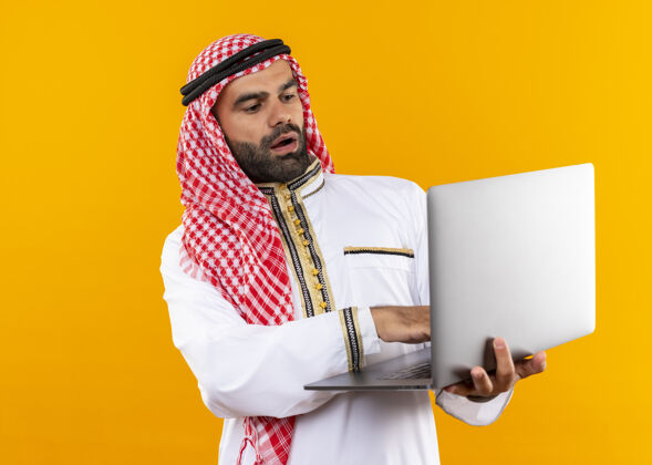 持有身着传统服装的阿拉伯商人手持笔记本电脑站在橙色的墙上 看上去既惊讶又惊奇穿笔记本电脑看
