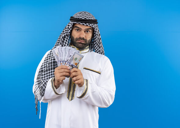 传统身着传统服装的阿拉伯男子站在蓝色的墙上 带着自信严肃的表情展示着现金认真站现金
