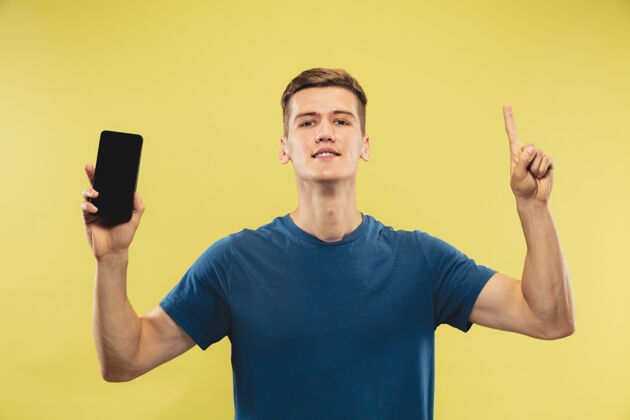 人黄色工作室背景上的白人年轻人半身肖像穿着蓝色衬衫的漂亮男模人类情感的概念 面部表情显示手机屏幕并指向上表情工作男性