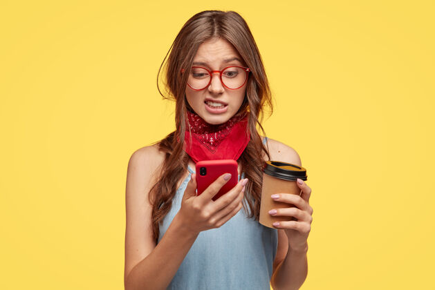 负片不满意的可爱女人在手机上厌恶地看着 在专门的应用程序里编辑照片 喝咖啡去 在黄色的墙上摆姿势 感到厌恶 连接到高速互联网短信眼镜白种人