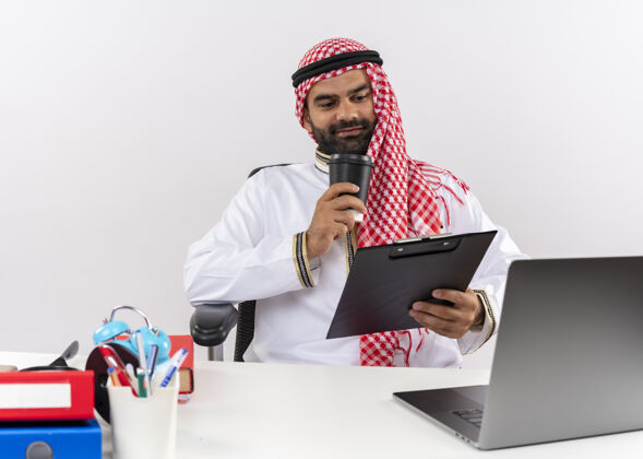电脑身着传统服装的阿拉伯商人坐在桌旁 手提电脑拿着剪贴板和咖啡杯 快乐而积极地在办公室工作阿拉伯语拿着正面