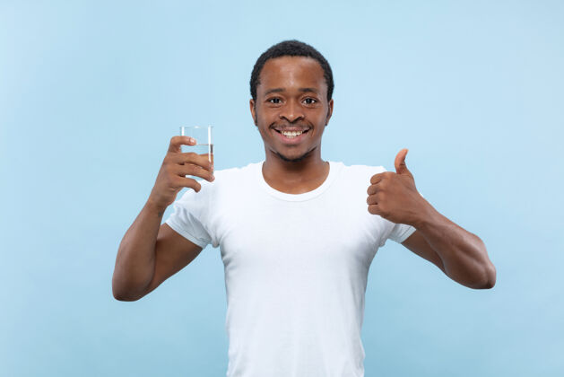 商人在蓝色背景上 一个身穿白衬衫的非洲裔美国年轻人的半身特写肖像人类的情感 面部表情 广告概念拿着一个玻璃杯和饮用水事业表情非洲