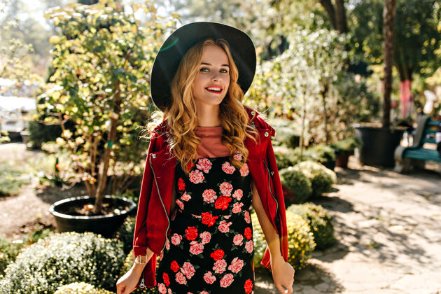 人迷人的卷发女子 戴着宽边帽 身着玫瑰裙 微笑着在公园摆姿势花店植物享受