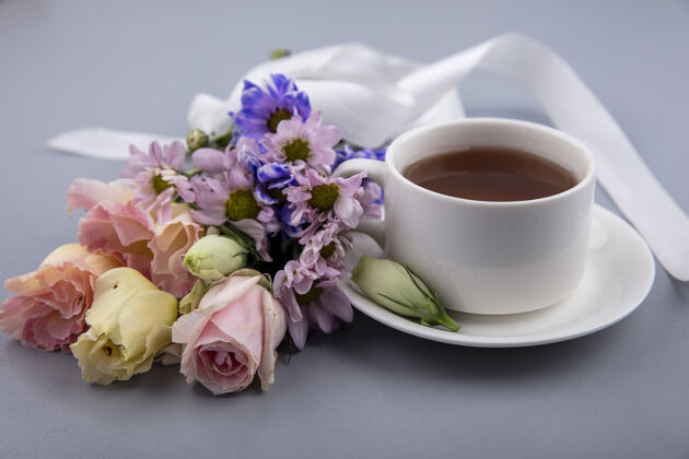 桌子茶碟上的茶杯和灰色背景上带有缎带的花朵的侧视图丝带茶碟饮料