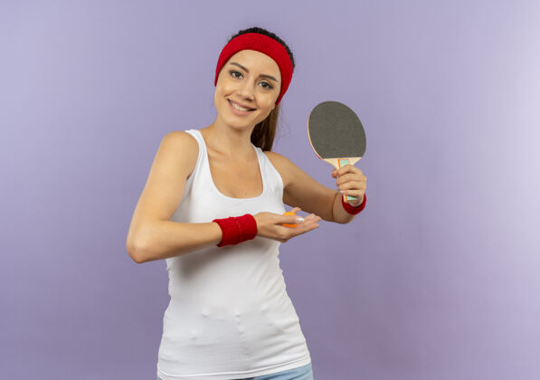 球穿着运动服的快乐的年轻健身女孩 戴着头带 拿着球拍和乒乓球 脸上带着微笑站在灰色的墙上欢呼脸站