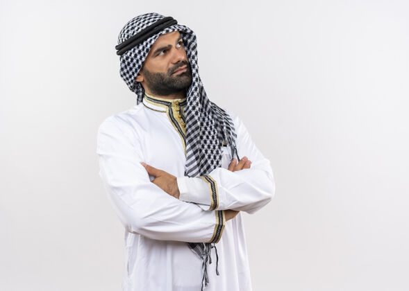 手臂身着传统服装的阿拉伯男人 双臂交叉放在胸前 严肃的脸站在白色的墙壁上向一边看传统站穿