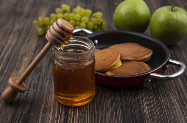 罐子前视图煎锅里的煎饼 蜂蜜放在木勺的罐子里 绿色葡萄和苹果放在木制背景上薯条苹果前面
