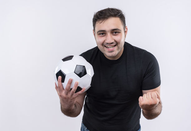 旅行身穿黑色t恤的年轻人手持足球棒握紧拳头站在白色的墙上 高兴而兴奋足球快乐市民