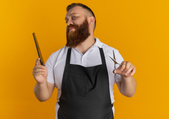 男人专业的胡须理发师站在橙色的墙上 围着围裙 手里拿着剪刀和发刷 脸上带着微笑胡子脸微笑