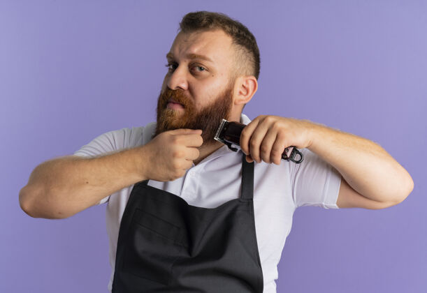 专业专业的胡须理发师站在紫色的墙上 围着围裙用剃须刀剪胡子 看上去很困惑困惑理发师男人