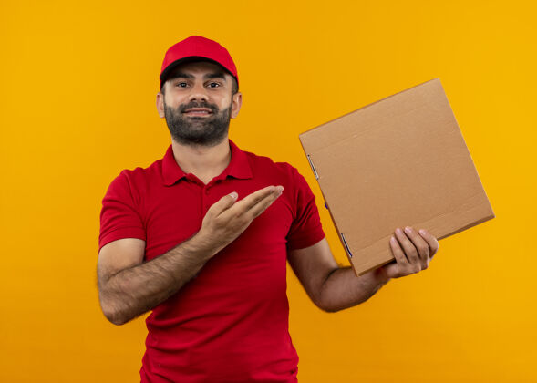 胡须满脸胡须 身穿红色制服 头戴鸭舌帽的送货员站在橘红色的墙上 手举披萨盒 自信地微笑着站立展示送货