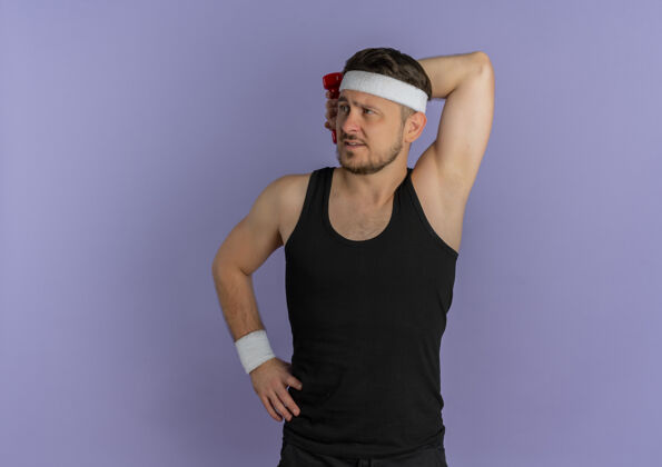 年轻戴着头巾的年轻健身男子一边看一边练习 一边用哑铃站在紫色的墙上站运动人
