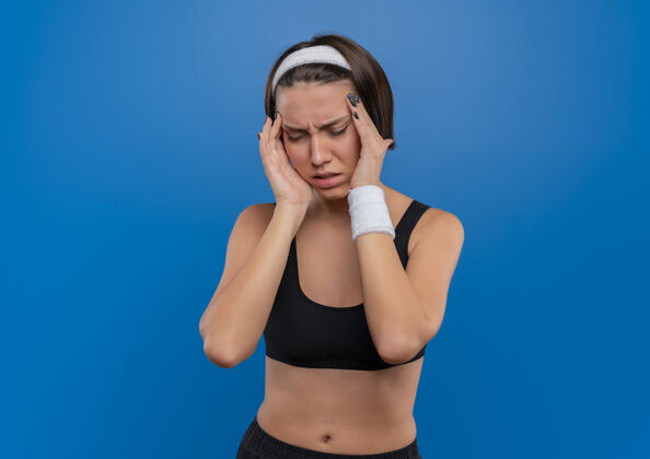 站立穿着运动服的年轻健身女士站在蓝色的墙上摸着太阳穴头痛女人太阳穴姿势
