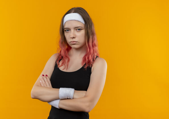 运动装身穿运动服 双臂交叉放在胸前 严肃的脸站在橙色的墙上的年轻健身女士运动健康健身