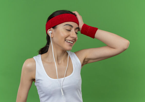 运动装身穿运动服 头戴头巾的年轻健身女士站在绿色的墙上 面带迷茫的微笑人市民微笑