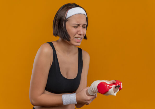 健身身穿运动服的年轻健身女 头戴哑铃 抚摸手腕 站在橘色的墙上 看起来不舒服 感觉疼痛市民手腕运动