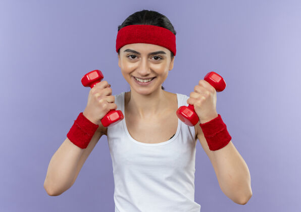 头带穿着运动服的年轻健身女士 头箍上举着两个哑铃欢呼举起健身