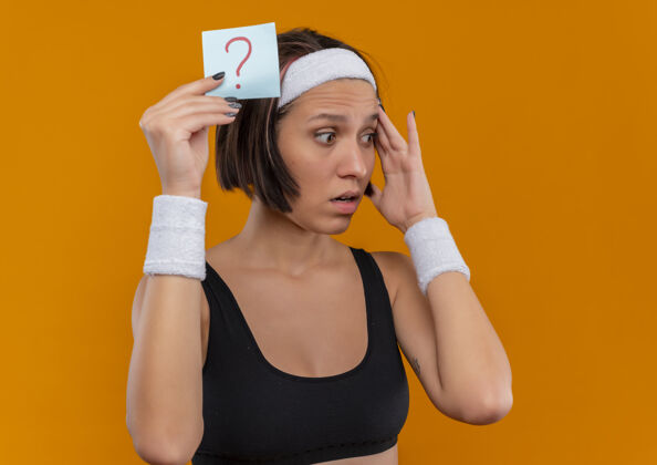 标记身穿运动服的年轻健身女 头上戴着头带 手里拿着带问号的提醒纸 站在橘色的墙上惊讶地看着一边女人旁边站着
