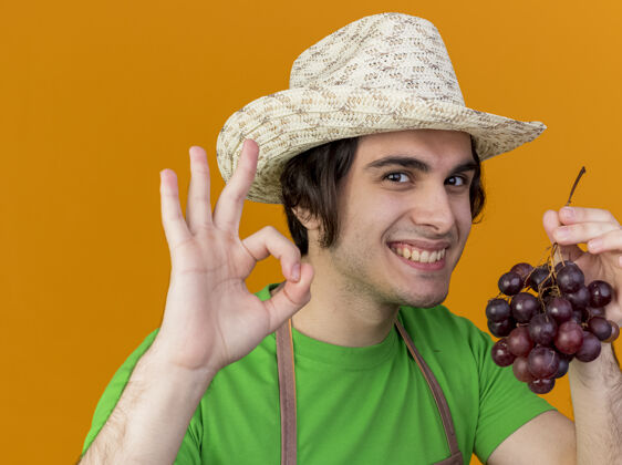 年轻年轻的园丁围裙戴着帽子 手里拿着一束葡萄 脸上挂着“ok”的牌子 站在橘色的背景上 开心地微笑着帽子微笑花园