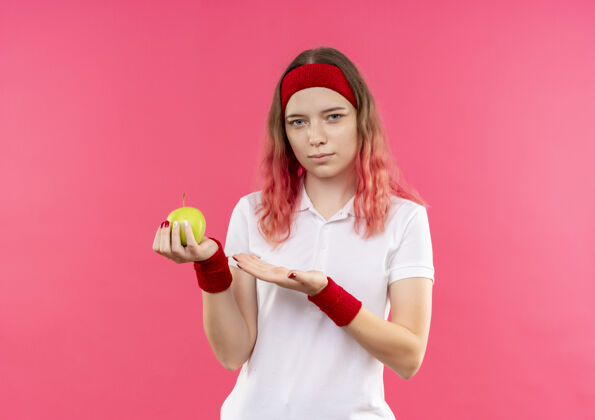 运动戴着头巾的年轻运动女性站在粉色的墙上 展示着绿苹果 她的手臂看起来很自信手人姿势