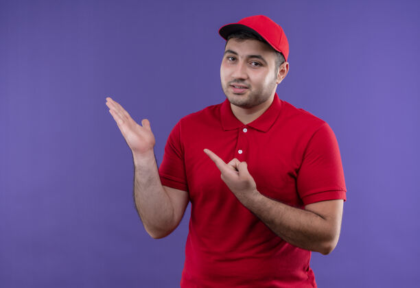 人身穿红色制服 戴着帽子的年轻送货员站在紫色的墙上 用手指着胳膊微笑着展示复印空间站立公民手