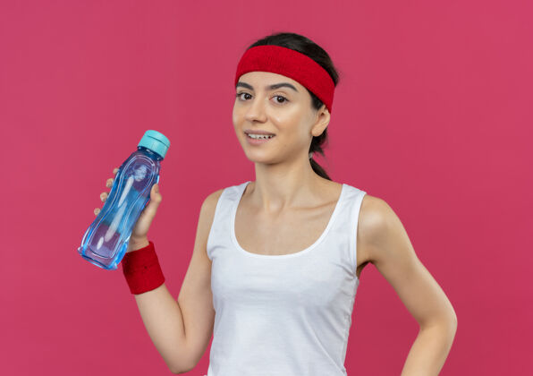 瓶子身穿运动服的年轻健身女 戴着头巾 手里拿着一瓶水 站在粉色的墙上 脸上带着微笑 看上去很自信抱人微笑