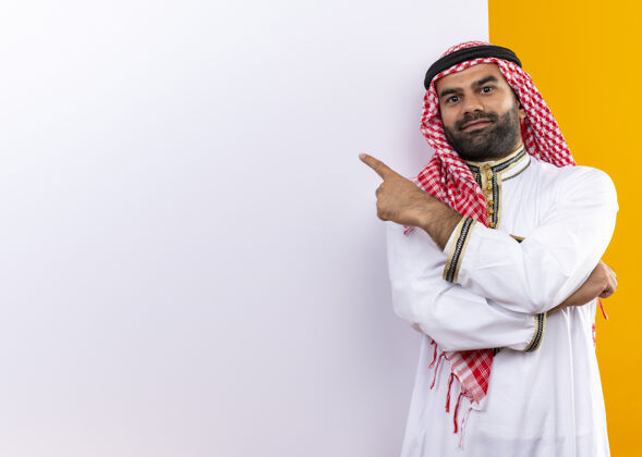 微笑身着传统服装的阿拉伯商人站在空白的广告牌旁 手指指着广告牌 脸上带着自信的微笑 越过橙色的墙壁阿拉伯语传统近