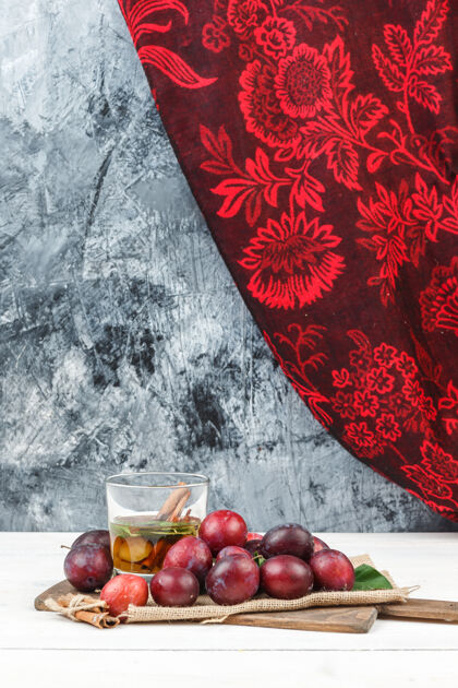 桌子用一个袋子把李子和排毒水放在切菜板上 白色木板和深蓝色大理石表面上有一个红色的窗帘垂直有机玻璃叶子