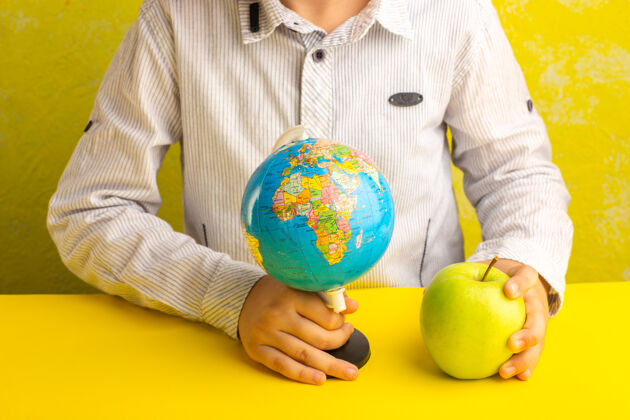 手持式正面图小男孩拿着小地球仪和绿色苹果在黄色的表面打击乐器苹果地球仪