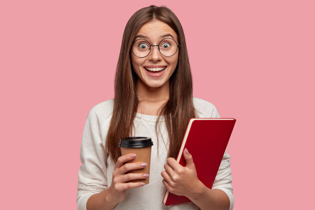快乐快乐的黑发女人表情兴奋 神情幸福 戴着圆眼镜 拿着红色课本 拿出咖啡 对同学的好消息做出反应 孤立在粉红色的墙上青少年茶满意