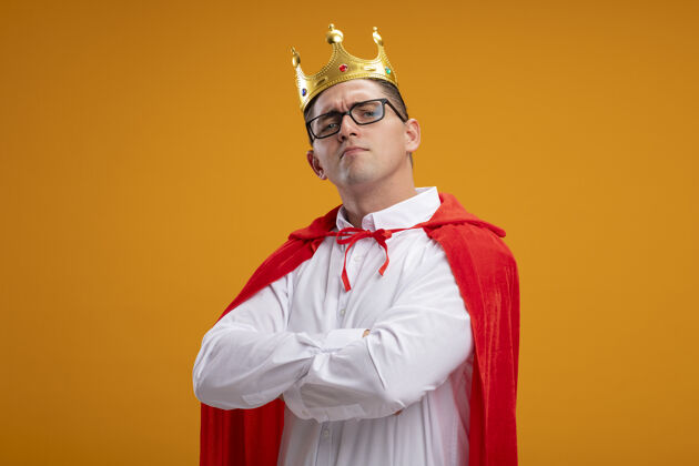 骄傲超级英雄商人穿着红色斗篷戴着眼镜戴着皇冠看着镜头感到骄傲自满胸前交叉双臂站在橙色背景之上皇冠英雄超级