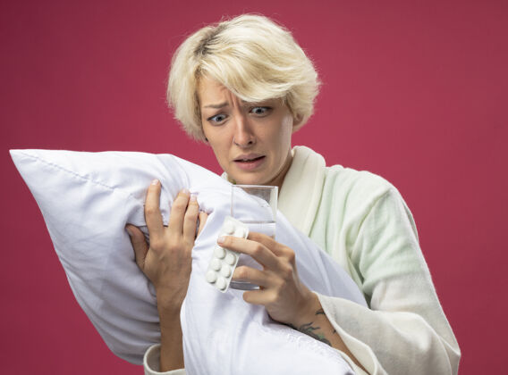 站着生病的不健康的短发妇女抱着枕头感觉不舒服拿着一杯水和药站在粉红色的背景下紧张不安感觉紧张药物