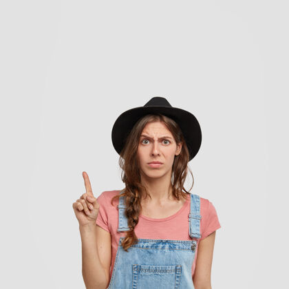 表情不高兴的女农场主的照片穿着休闲工作服 戴着时髦的黑帽子 指着上角 对新买的东西不满意 表示不喜欢 注意到一些无用的东西恼怒成人负面
